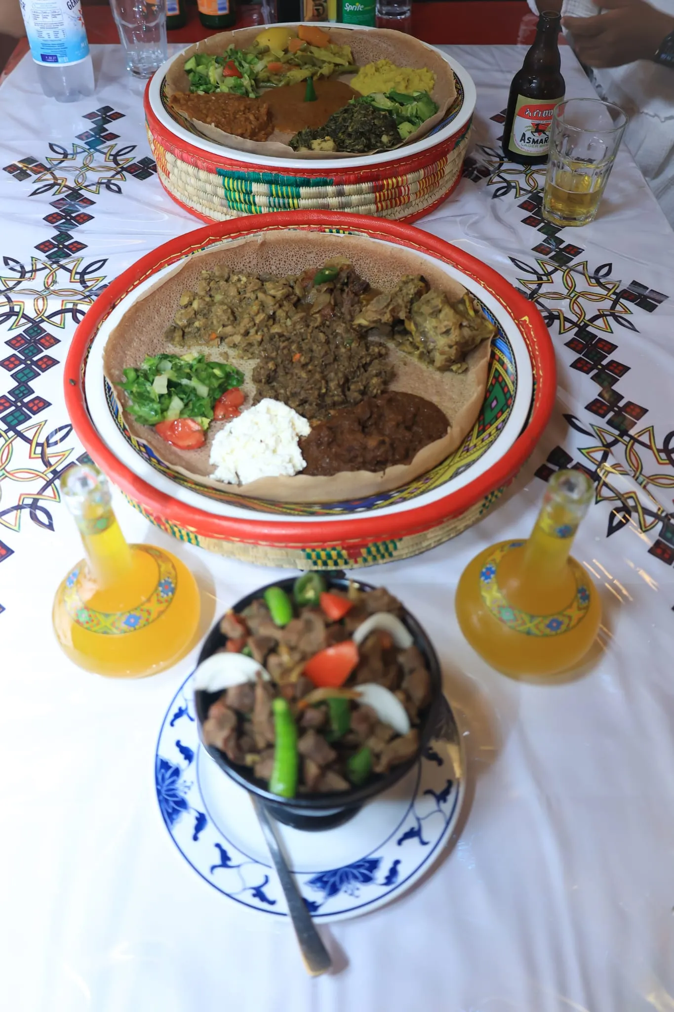 Gedeckter Tisch bei Bahgna: zwei Injera-Platten in roten Körben mit Fleisch- und Gemüsetöpfen, Tibs im schwarzen Tontopf und zwei Karaffen goldener Tej