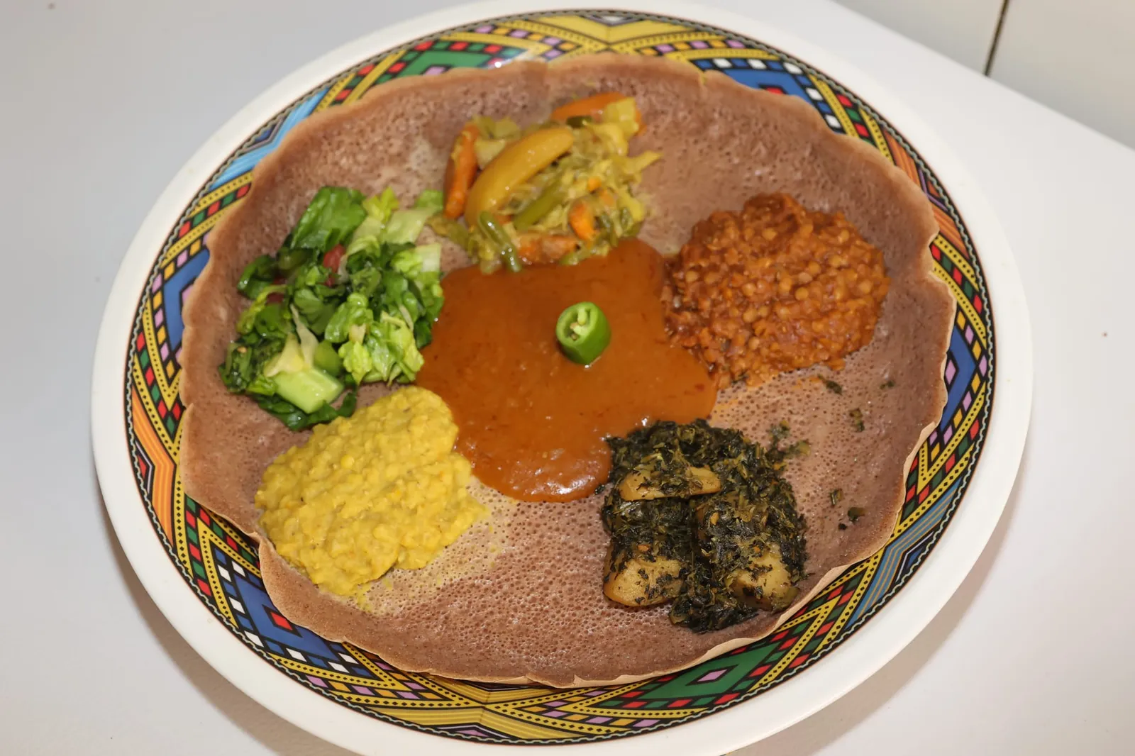 Injera-Platte mit buntem Regenbogen-Rand: Berbere-Sauce mit grüner Chili in der Mitte, ringsum Gemüse, Salat, rote Linsen und gelbes Erbsenpüree